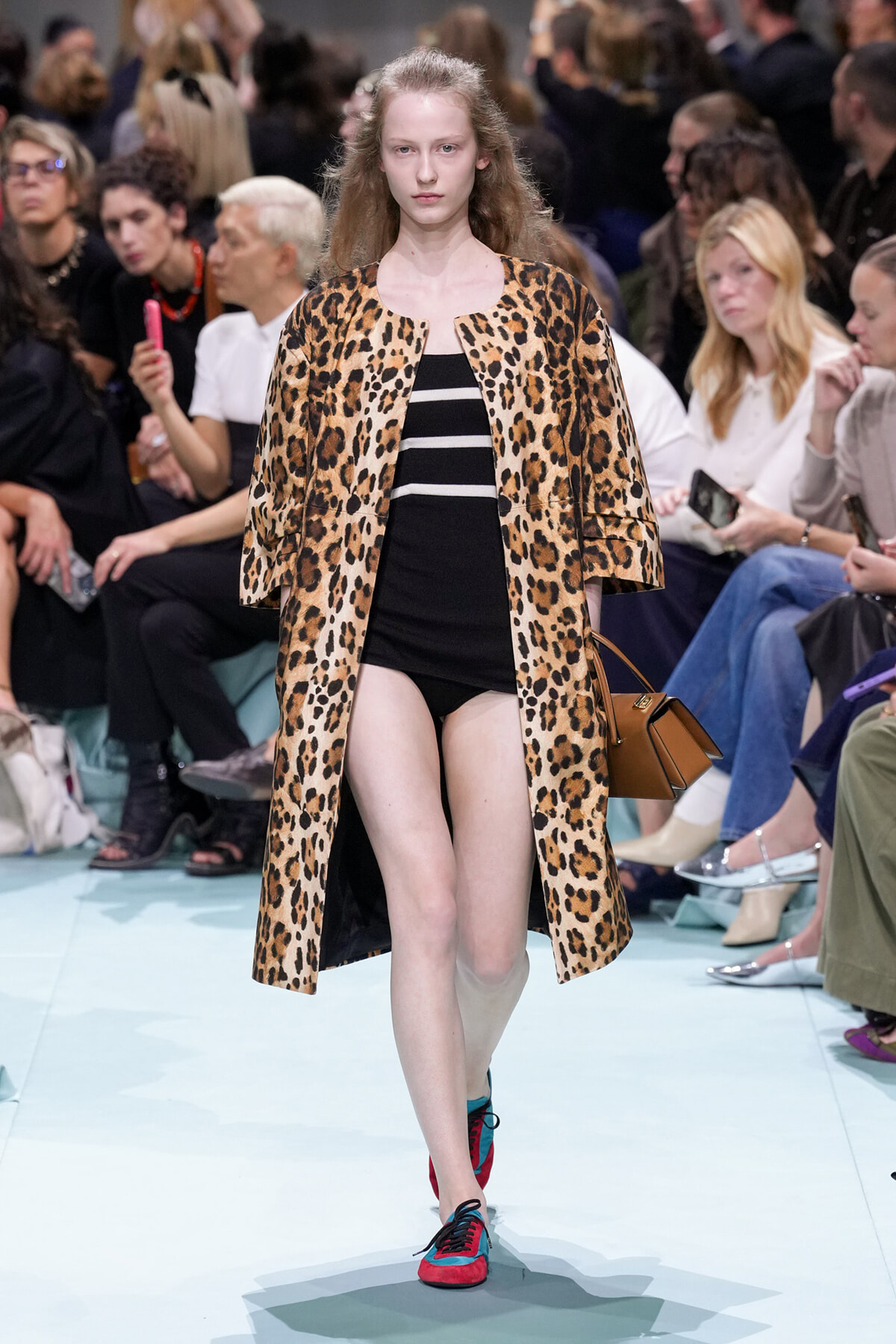 Model walks the runway in a leopard-print coat over a black striped mini dress, with a brown handbag at her side, audience in background.