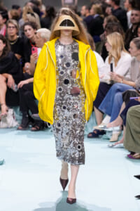 Model on a fashion show runway wearing a metallic silver sequin dress with circular cutouts, a bright yellow windbreaker, and a beige sculptural visor helmet obscuring the face, paired with burgundy pointed-toe heels.