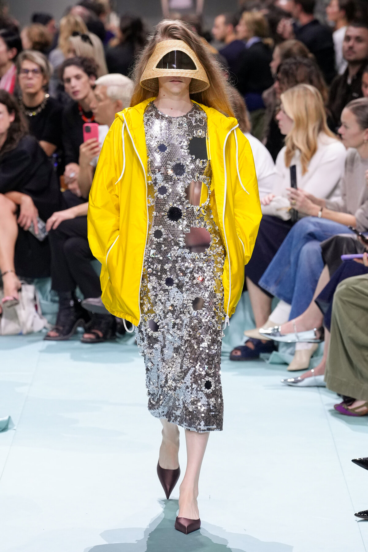 Model on a fashion show runway wearing a metallic silver sequin dress with circular cutouts, a bright yellow windbreaker, and a beige sculptural visor helmet obscuring the face, paired with burgundy pointed-toe heels.