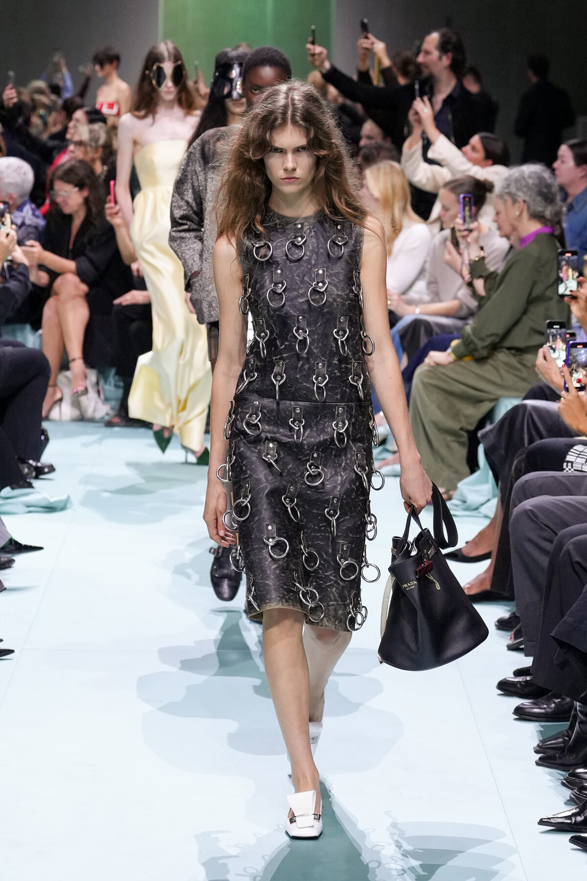 Model walking a light-blue runway in a black textured dress adorned with metal rings, carrying a black handbag.
