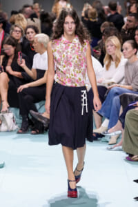 Model walks a fashion show runway in a pink floral sleeveless blouse and navy skirt with white lace-up detail, surrounded by seated audience members.