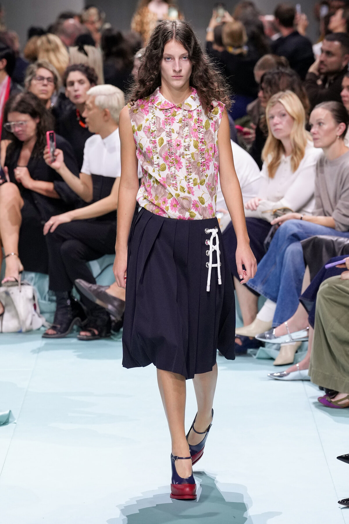 Model walks a fashion show runway in a pink floral sleeveless blouse and navy skirt with white lace-up detail, surrounded by seated audience members.
