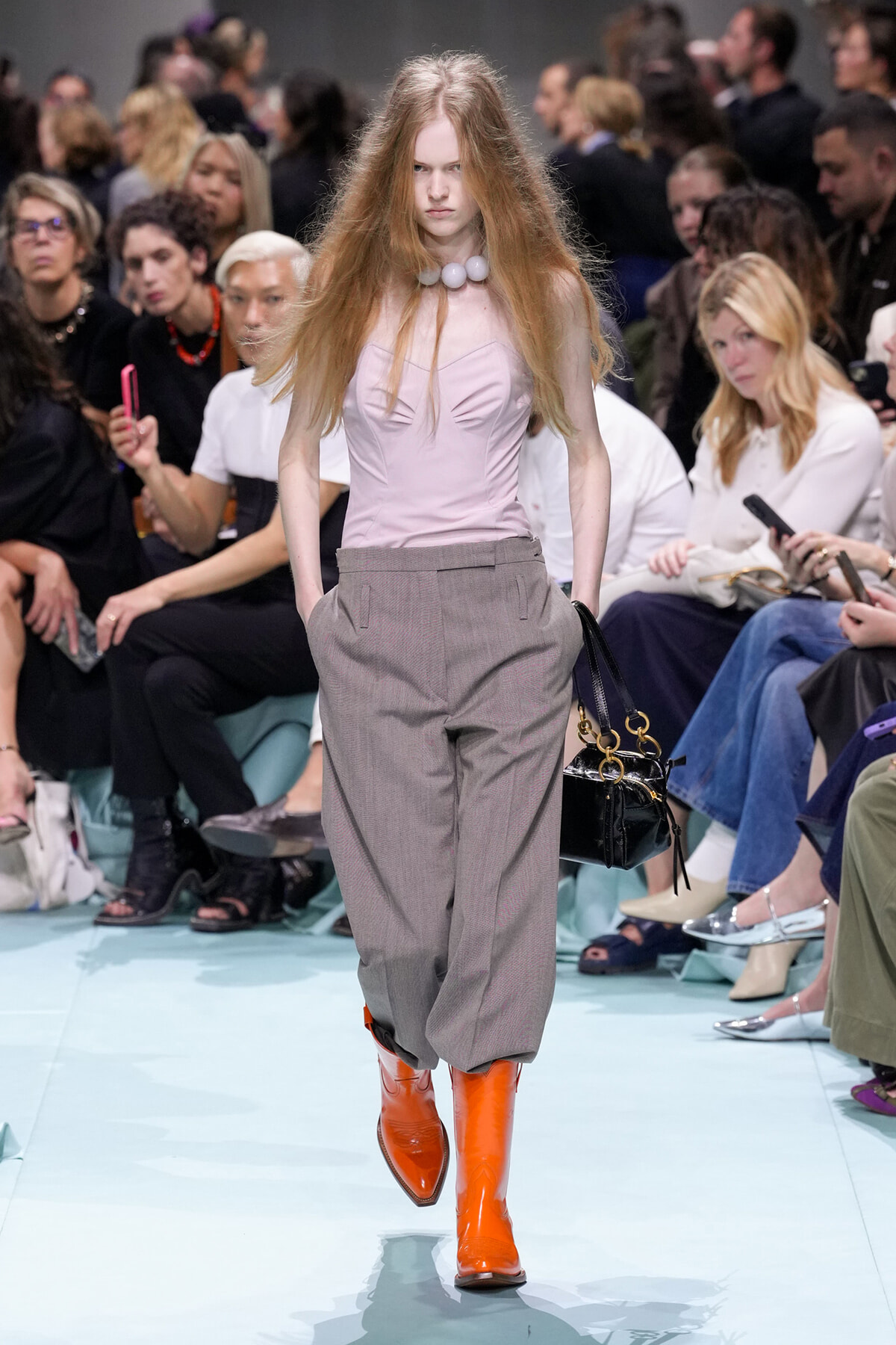 Model walks a fashion runway wearing a pale pink corset-style top, loose grey trousers, and bright orange knee-high boots; audience watches in Background, holding phones.