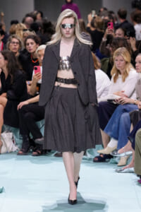 Model walking a runway in a dark gray blazer and pleated skirt, metallic chest armor, silver visor sunglasses, and a wide belt.