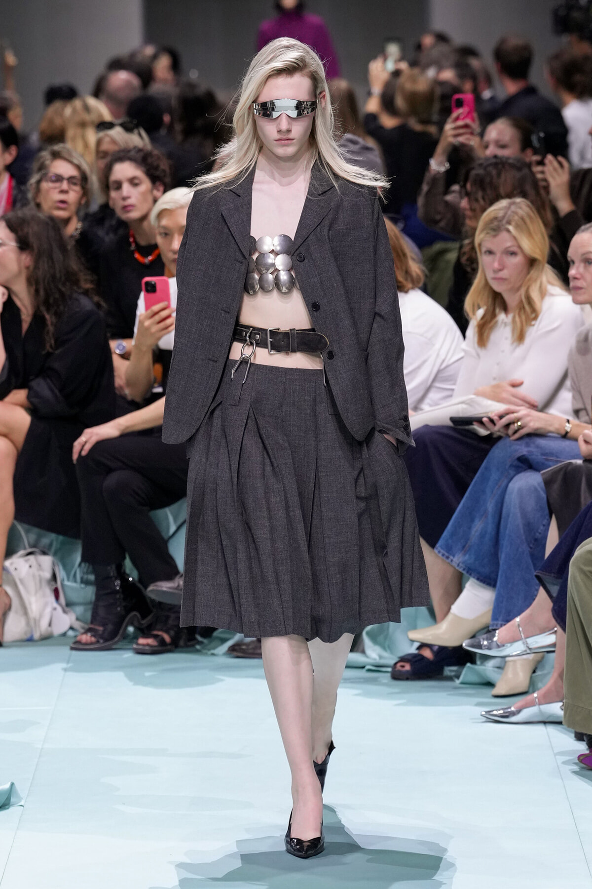Model walking a runway in a dark gray blazer and pleated skirt, metallic chest armor, silver visor sunglasses, and a wide belt.