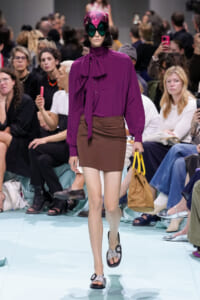 Model walks the runway in a bold purple blouse with a neck bow, brown mini skirt, and oversized pink visor sunglasses, carrying a tan bag with yellow handles.