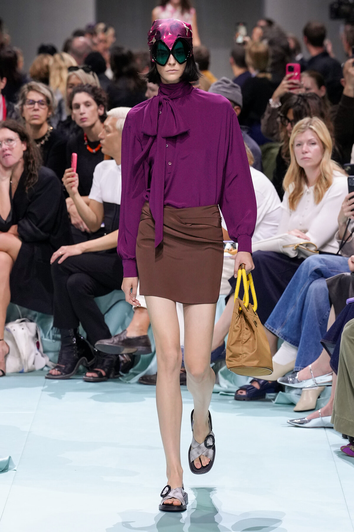 Model walks the runway in a bold purple blouse with a neck bow, brown mini skirt, and oversized pink visor sunglasses, carrying a tan bag with yellow handles.