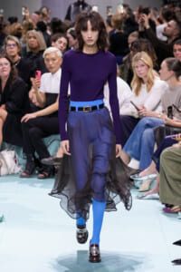 Model walks a runway in a royal purple knit top, electric blue belt, and blue trousers with a black sheer overlay flowing around the legs.