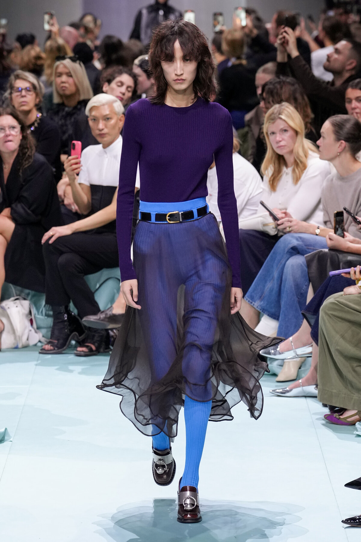 Model walks a runway in a royal purple knit top, electric blue belt, and blue trousers with a black sheer overlay flowing around the legs.
