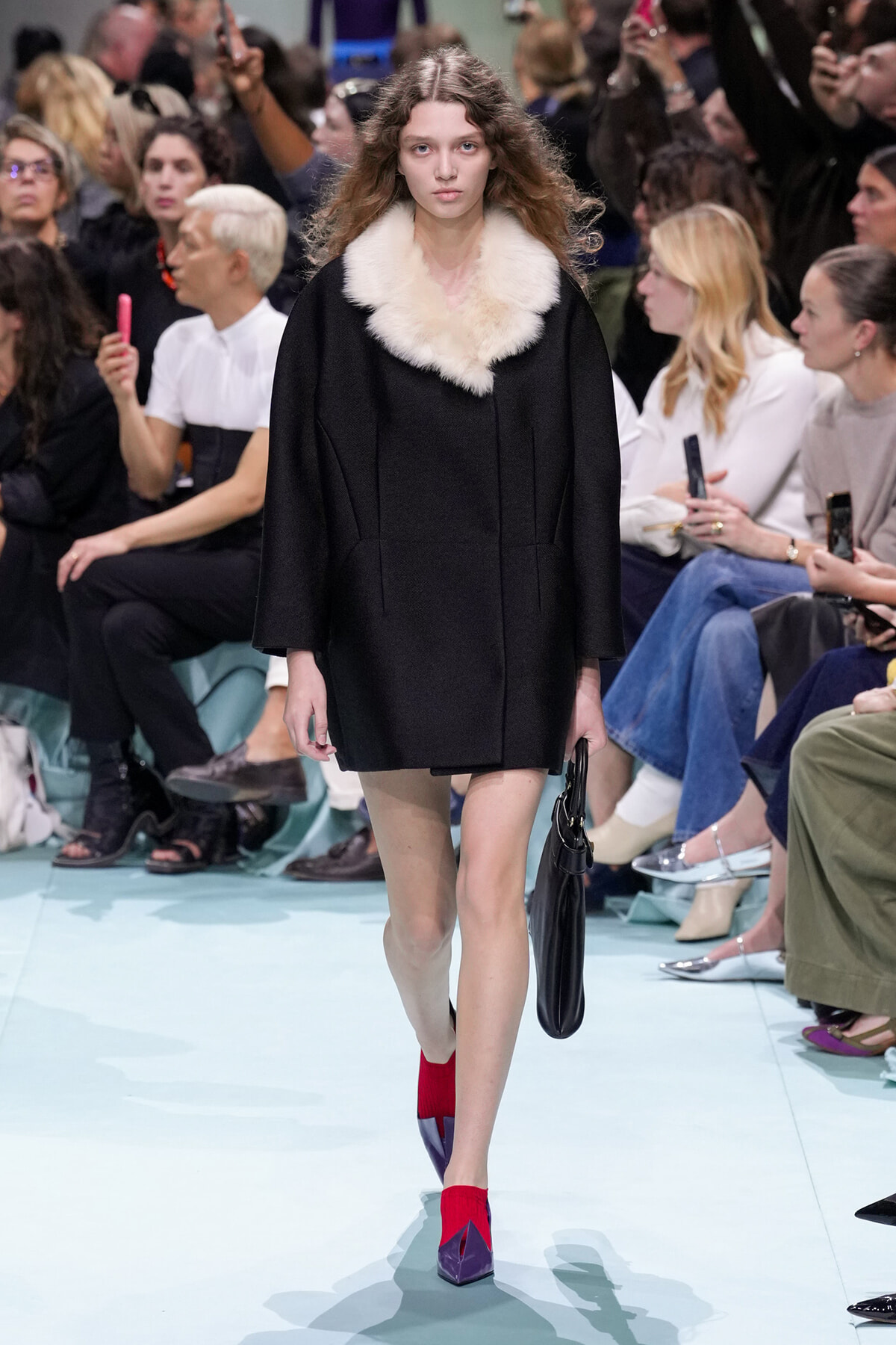 Model walking a runway in a black coat with a large cream fur collar, carrying a black handbag, and wearing red socks with purple heels as spectators watch.