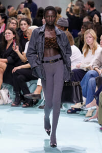 Runway model wearing a gray jacket over a brown textured bustier, with gray ribbed leggings, a belt, and a black handbag on a fashion show runway.