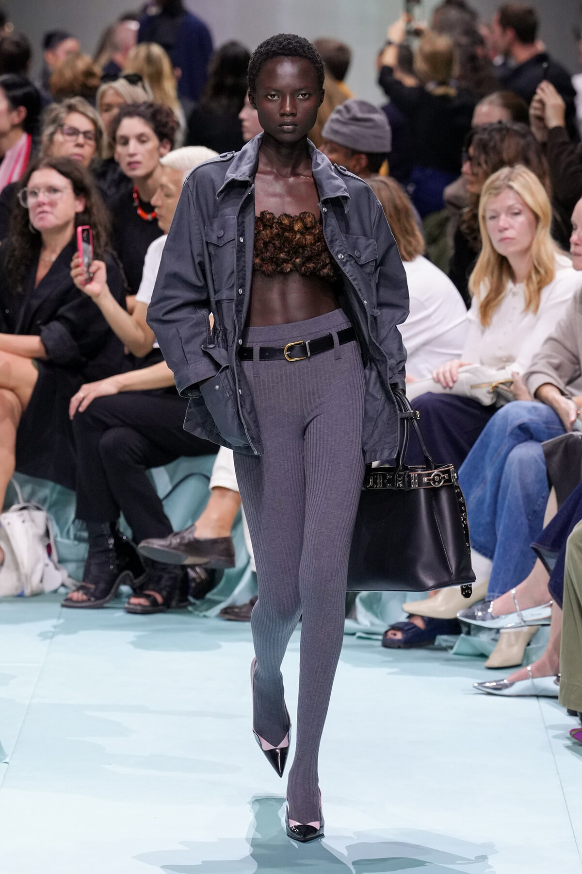 Runway model wearing a gray jacket over a brown textured bustier, with gray ribbed leggings, a belt, and a black handbag on a fashion show runway.