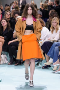 Model walking runway in an orange A-line skirt and camel coat with pink collar, black bralette visible underneath, crowd in background.