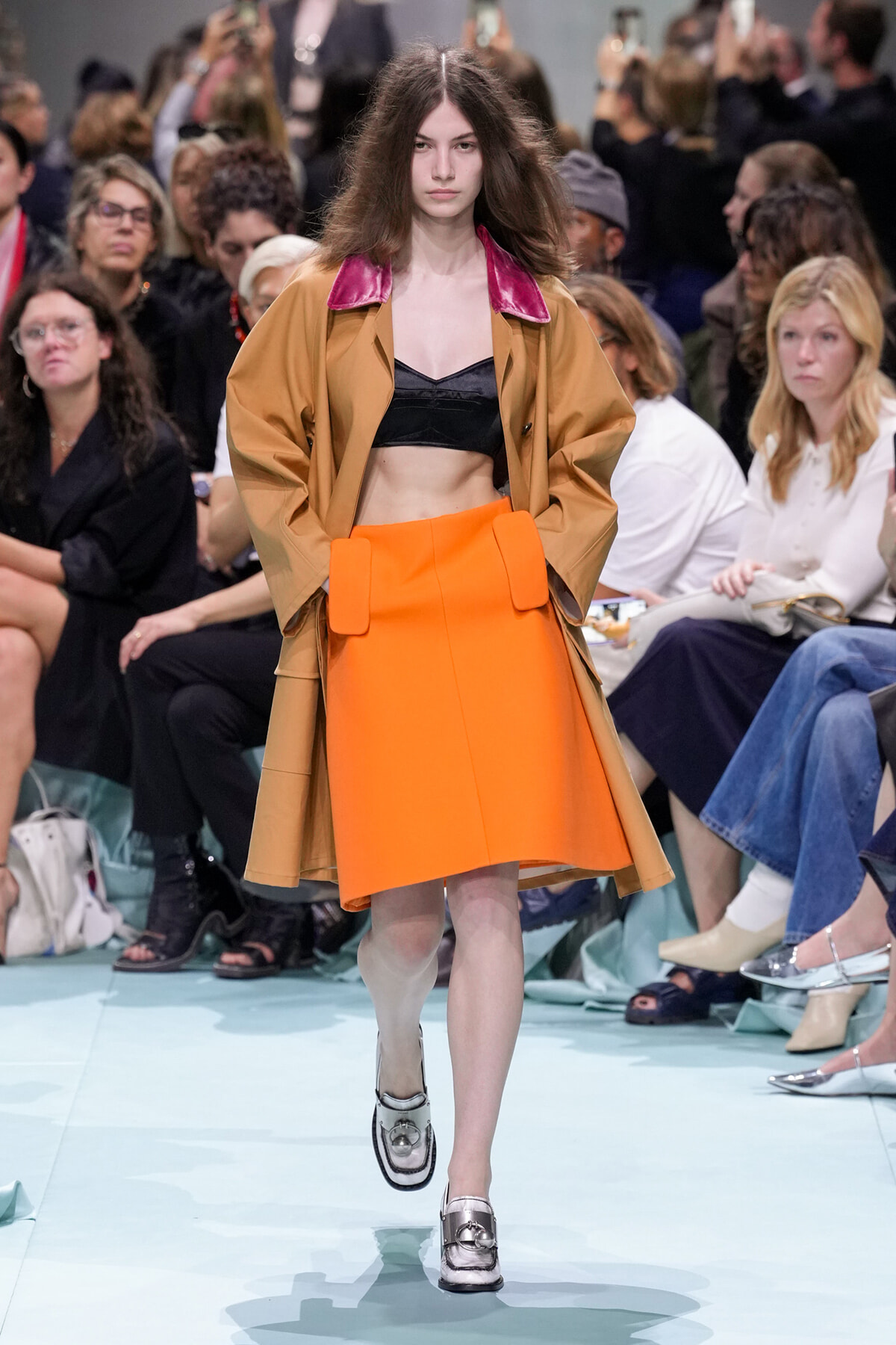 Model walking runway in an orange A-line skirt and camel coat with pink collar, black bralette visible underneath, crowd in background.