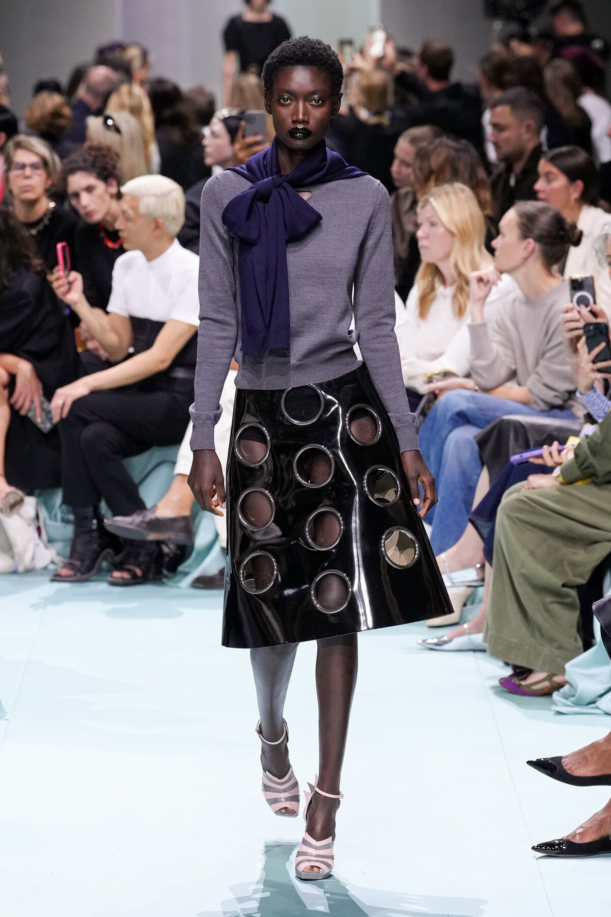 Runway model with short hair in a gray knit top, navy scarf, and glossy black circle-cutout A-line skirt on a light blue catwalkchic look.