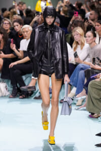 Model walking the runway in a shiny black leather coat with hood, black shorts, bright yellow high-heeled sandals, and a lavender handbag as spectators watch.