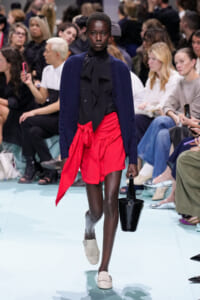 Model walking runway in navy cardigan over black blouse with bow, bright red wrap skirt, and beige loafers, carrying a black handbag; audience in background