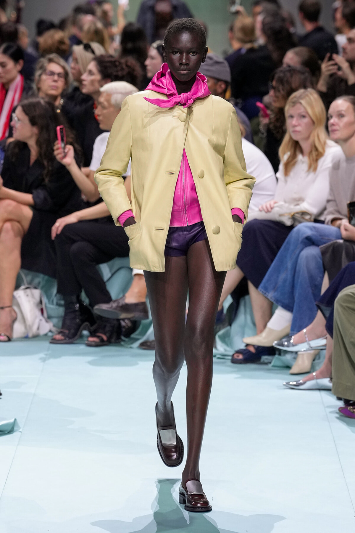 Runway model walks in a pale yellow coat over a bright pink top and scarf, with purple shorts and a gray prosthetic leg on the left.
