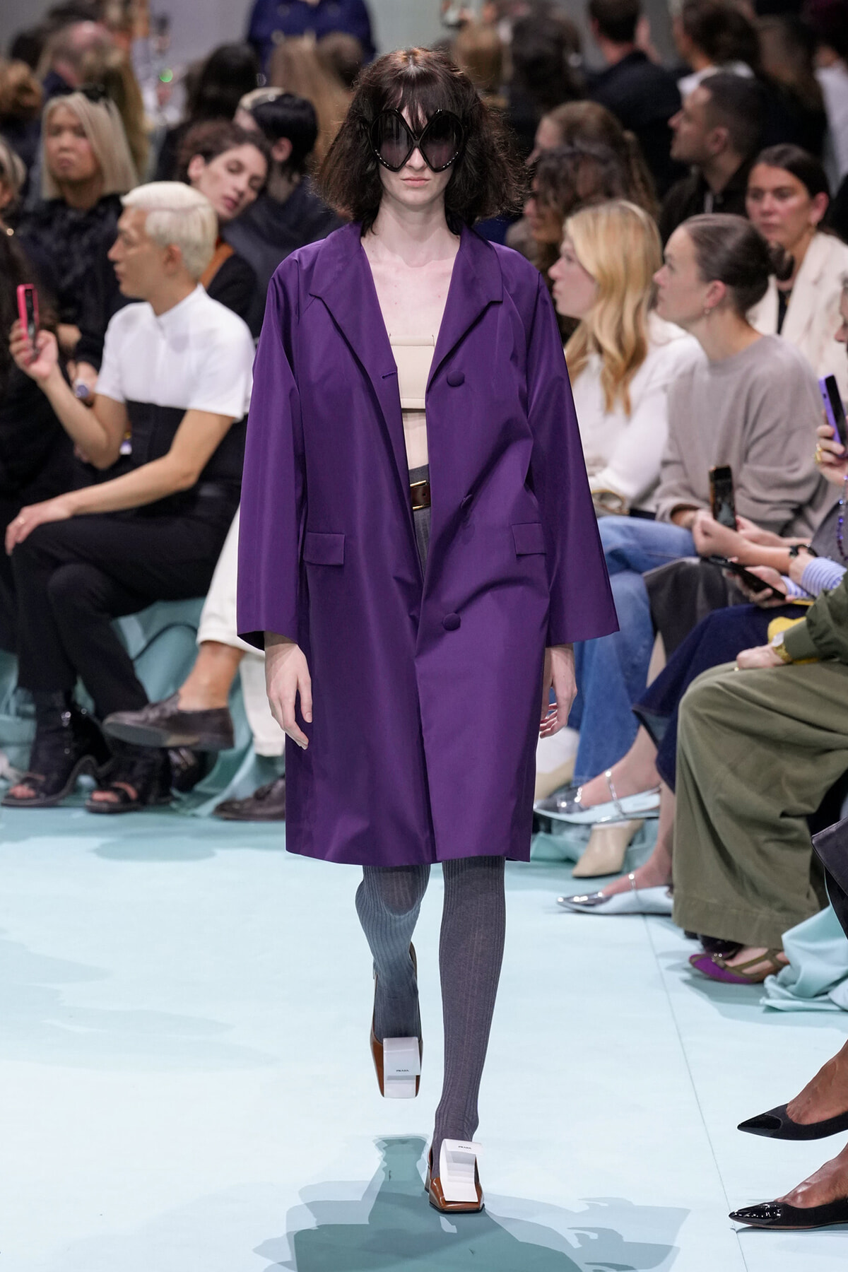 Model walks a fashion runway in a loose purple coat over a nude top, gray tights, and white platform shoes with oversized black sunglasses; audience watches