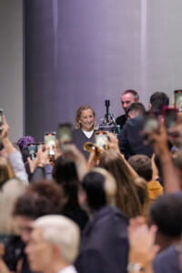 Smiling woman on stage surrounded by a crowd filming with smartphones at a public event.