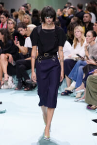 Model walks the runway in a black top and navy midi skirt with a statement belt and metal rings; audience watches closely.