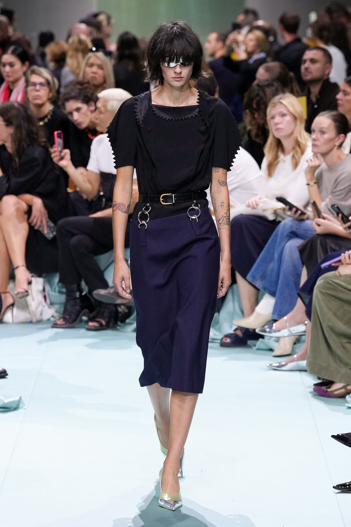 Model walks the runway in a black top and navy midi skirt with a statement belt and metal rings; audience watches closely.