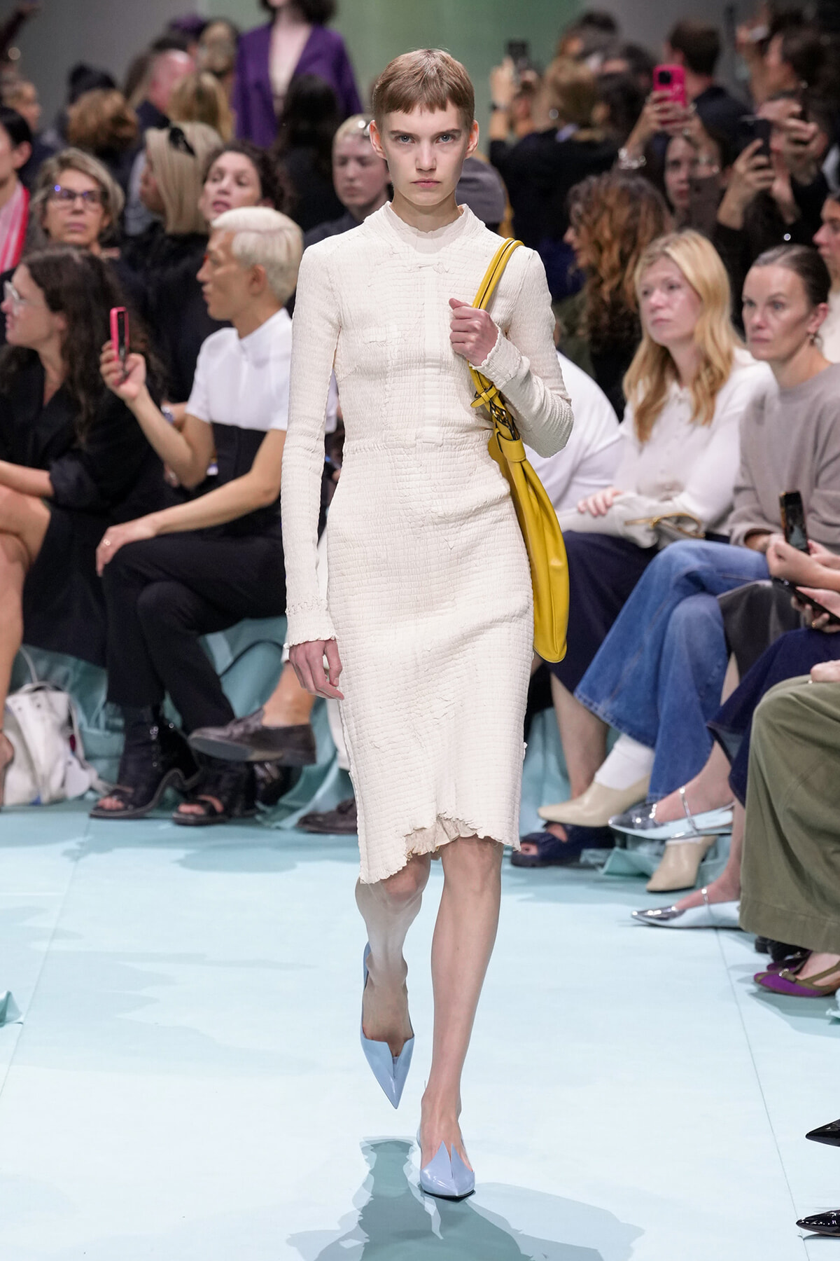 Model walks a fashion runway in a cream textured midi dress, bright yellow handbag over her shoulder, and light blue pointed heels as the crowd watches.