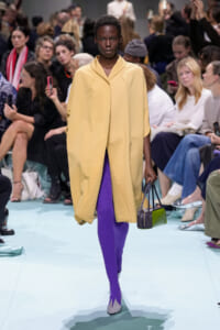Model on a runway wearing a pale yellow, oversized coat, bright purple ribbed tights, lavender heels, and a small two-tone green/purple handbag.