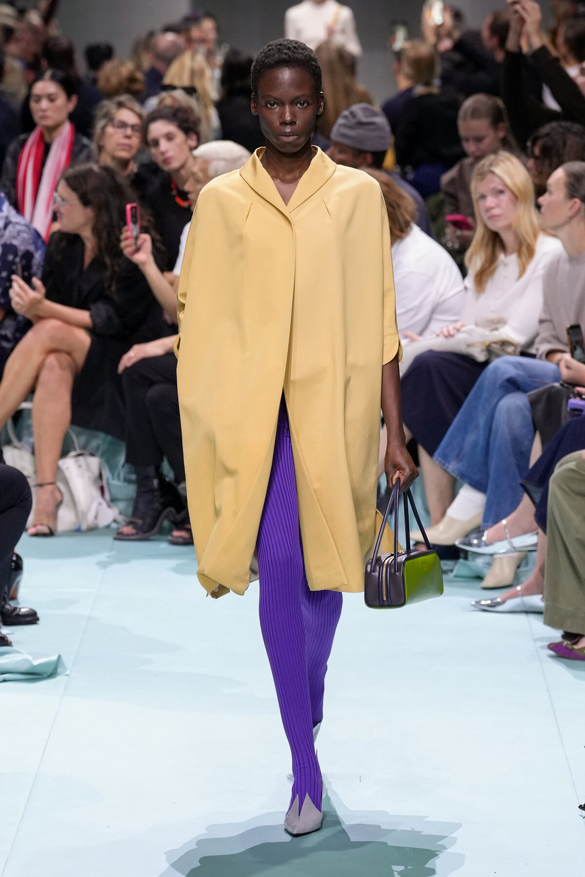 Model on a runway wearing a pale yellow, oversized coat, bright purple ribbed tights, lavender heels, and a small two-tone green/purple handbag.