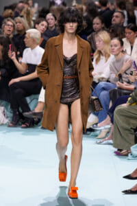 Model walks a fashion runway in a brown suede coat over a dark leather romper and bright orange platform heels, audience watching.