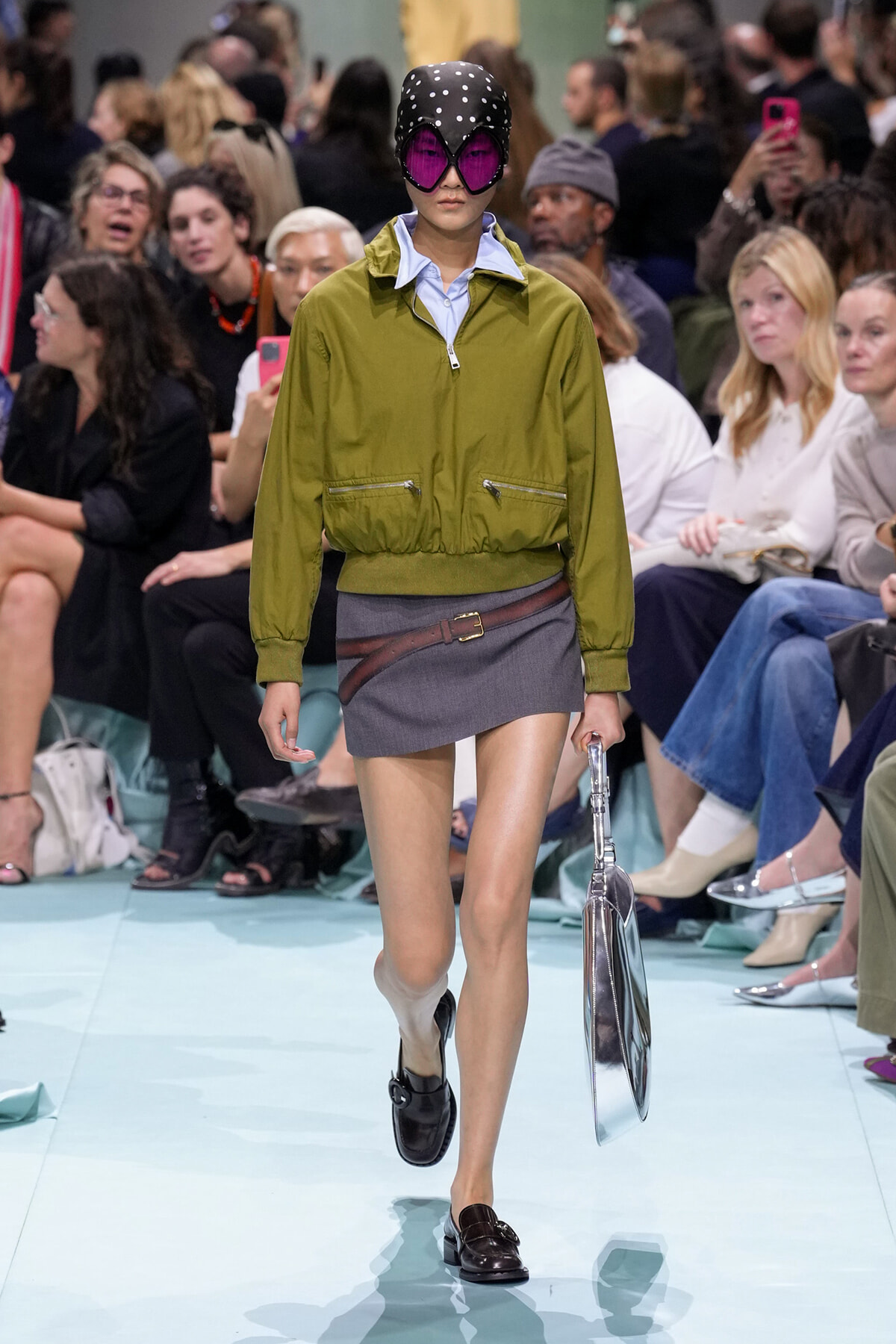 Model walks a fashion runway wearing an olive cropped jacket over a light blue shirt, gray miniskirt with brown belt, black loafers, and a polka-dot cap with pink sunglasses; holding a metallic handbag.