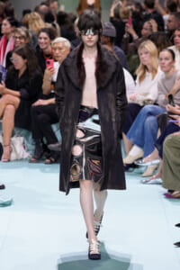 Model walking the runway in a black coat with a fur collar, paired with a shiny metallic skirt featuring circular cutouts and futuristic visor sunglasses in front of a seated audience.