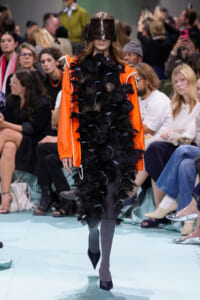 Model on a runway wearing an orange jacket over a black feathered dress, with a dark leather visor hat and gray tights, black heels.