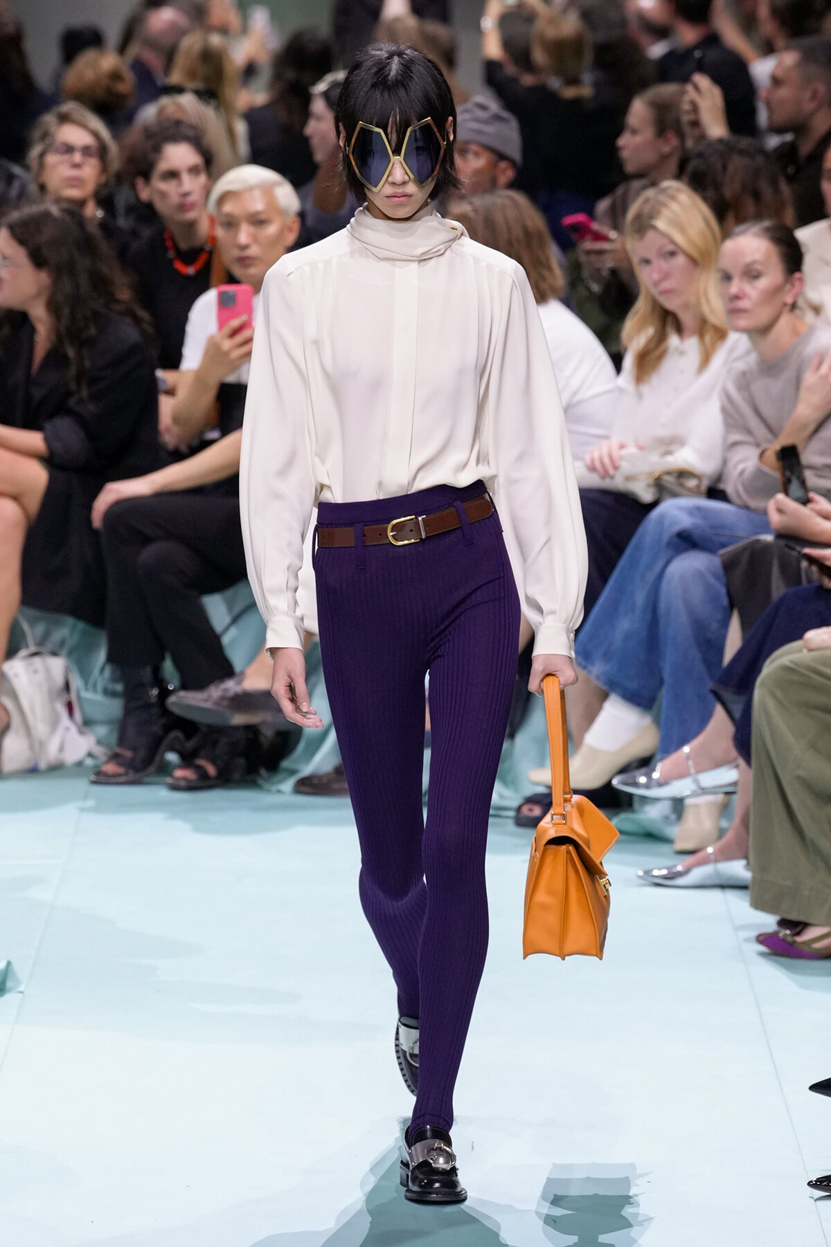 Model struts the runway in a cream blouse, dark purple ribbed tights, a brown belt, black loafers, and a bright orange handbag, wearing oversized geometric sunglasses.