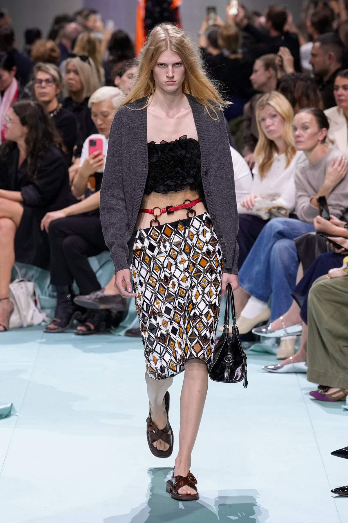 Runway model in a gray cardigan over a black ruffled strapless top, a geometric patterned skirt with orange accents, red belt, and brown sandals, carrying a black bag; audience in background.