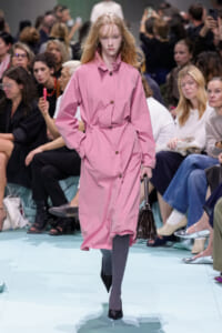 Model walking the runway in a pink belted trench coat with brown buttons, hands in pockets, carrying a black handbag while an audience watches.