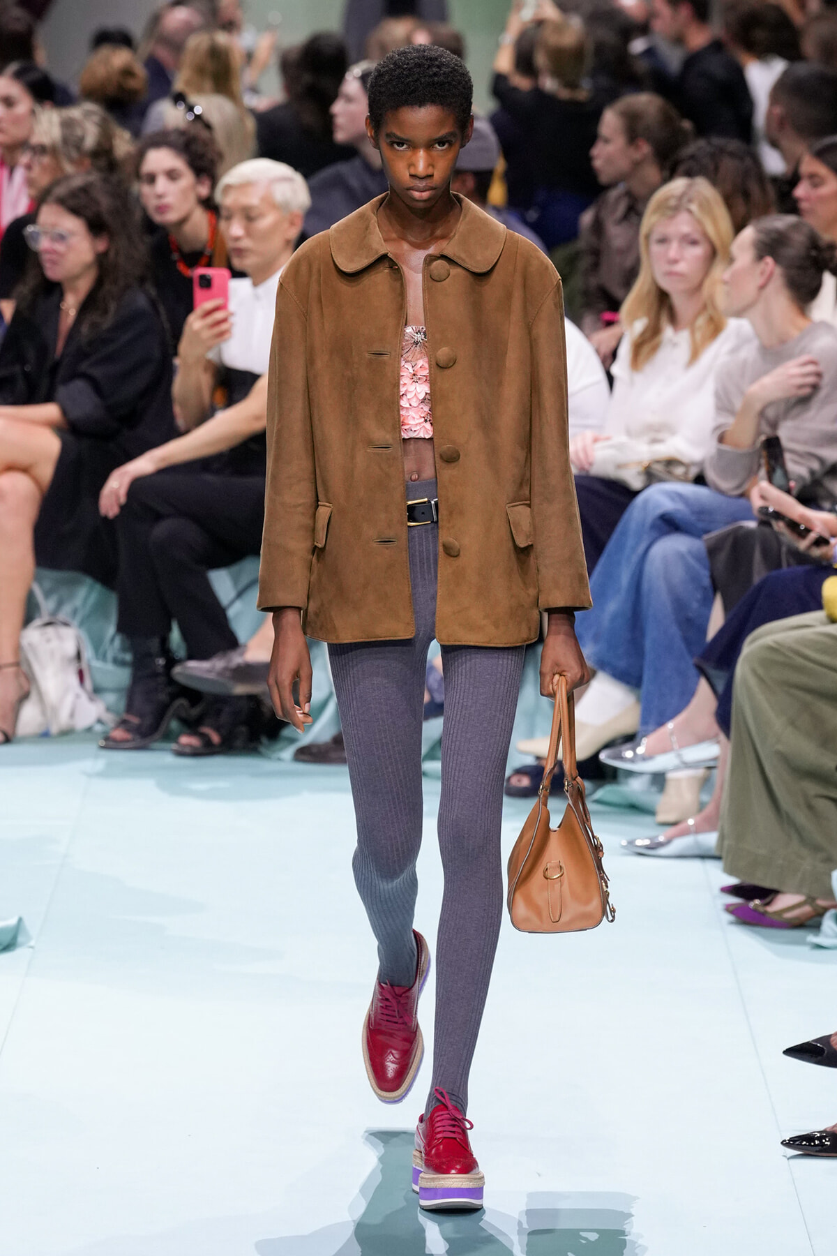 Model walks the runway in a brown suede jacket over a pink top, gray tights, and red platform shoes, carrying a tan handbag.