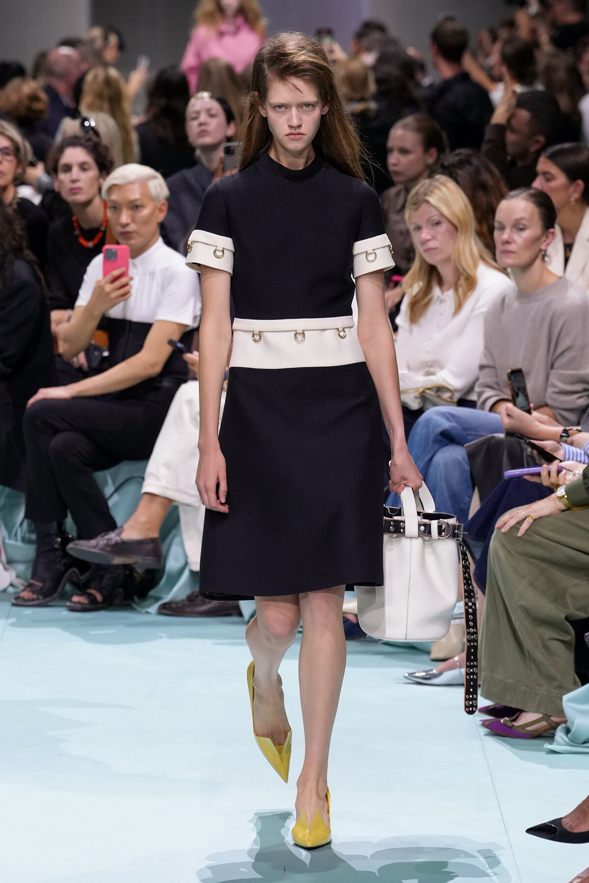 Model walks a runway in a black dress with white belts and yellow pointed heels, carrying a white handbag; audience watches in the background.