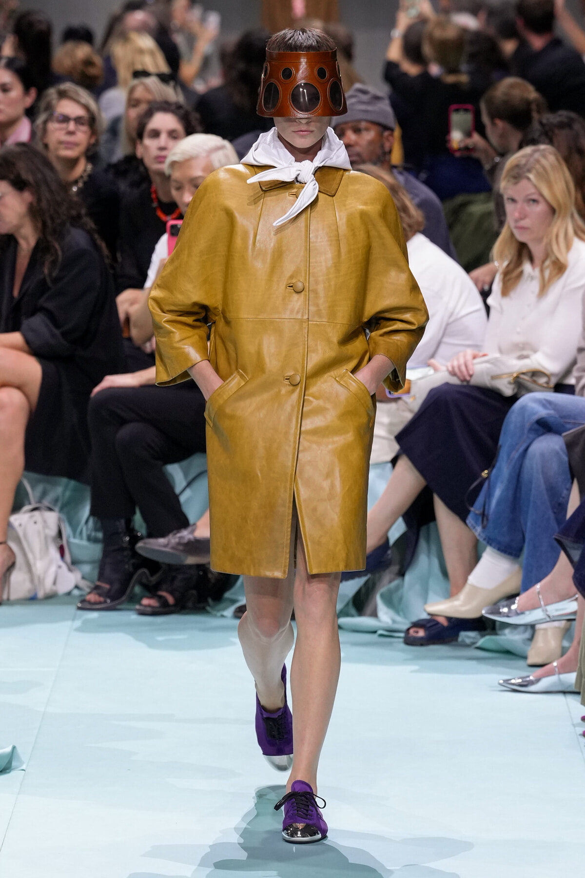 Model on a runway wearing a mustard leather coat with hands in pockets, white scarf, and a brown perforated helmet that covers the eyes; purple shoes and a light blue runway floor.
