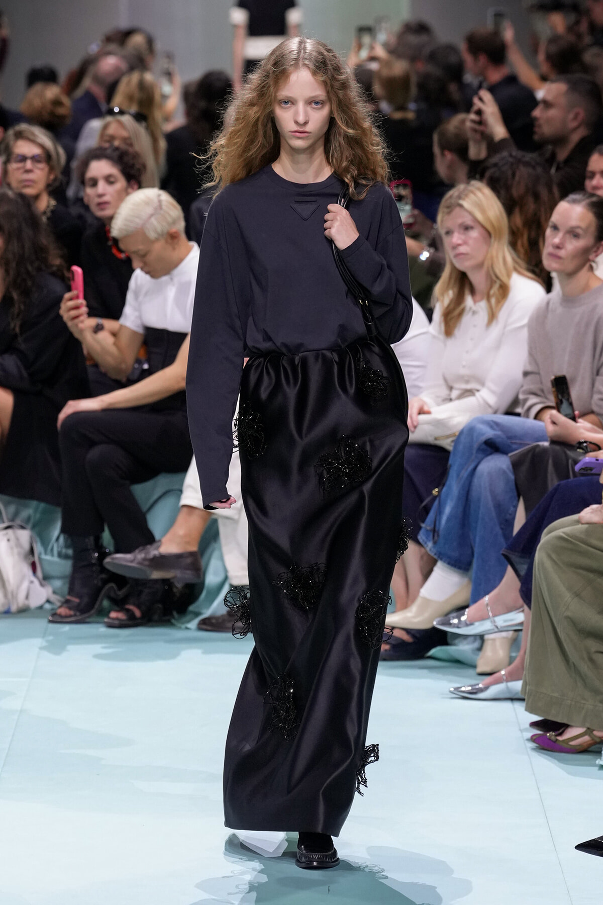 Model walking a fashion runway in a dark navy top and black satin maxi skirt with black floral appliqués; audience seated and taking photos in the background.