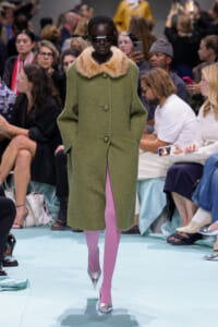 Model walks the runway in an olive green coat with fur collar, pink tights, and metallic silver heels, surrounded by an audience.