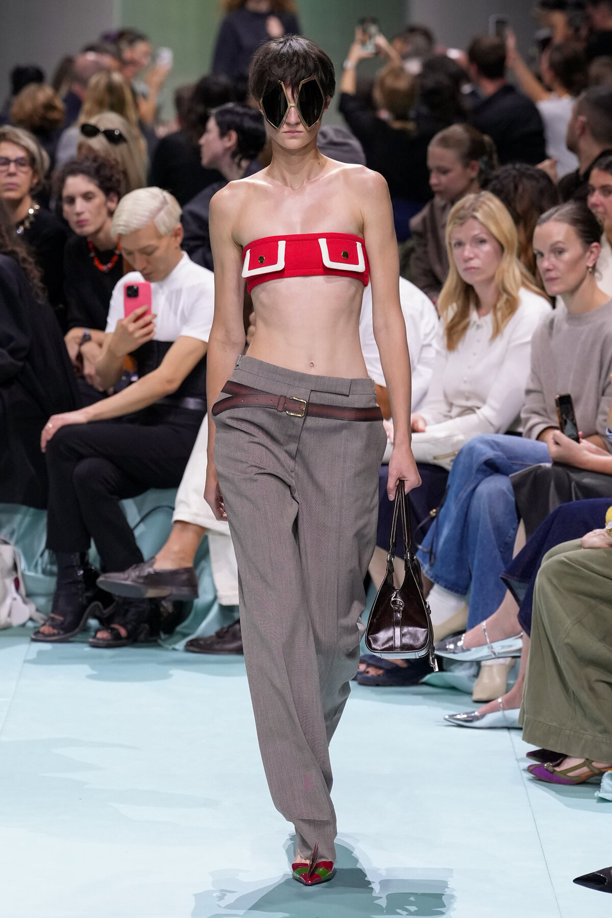 Model on a fashion runway wearing a red bandeau top, gray tailored pants, oversized sunglasses, and carrying a dark handbag.
