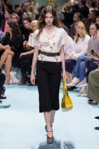 Model on a fashion runway wearing a cream bow-neck blouse, black pleated culottes, a chunky belt with metal rings, and brown-strap shoes; holding a yellow handbag. Audience in background.