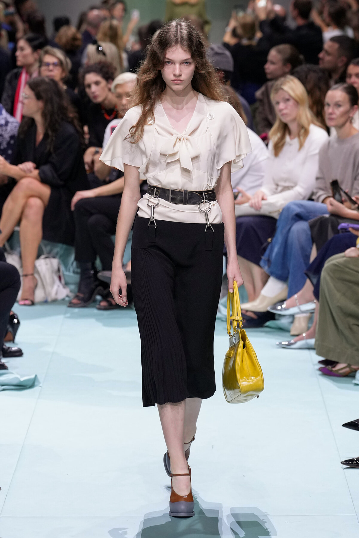 Model on a fashion runway wearing a cream bow-neck blouse, black pleated culottes, a chunky belt with metal rings, and brown-strap shoes; holding a yellow handbag. Audience in background.