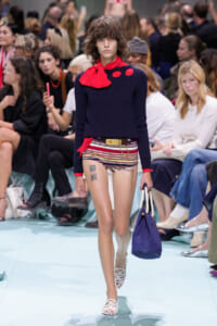 Model walks a fashion runway in a navy knit top with a red scarf, striped shorts, and a gold belt, carrying a purple handbag.