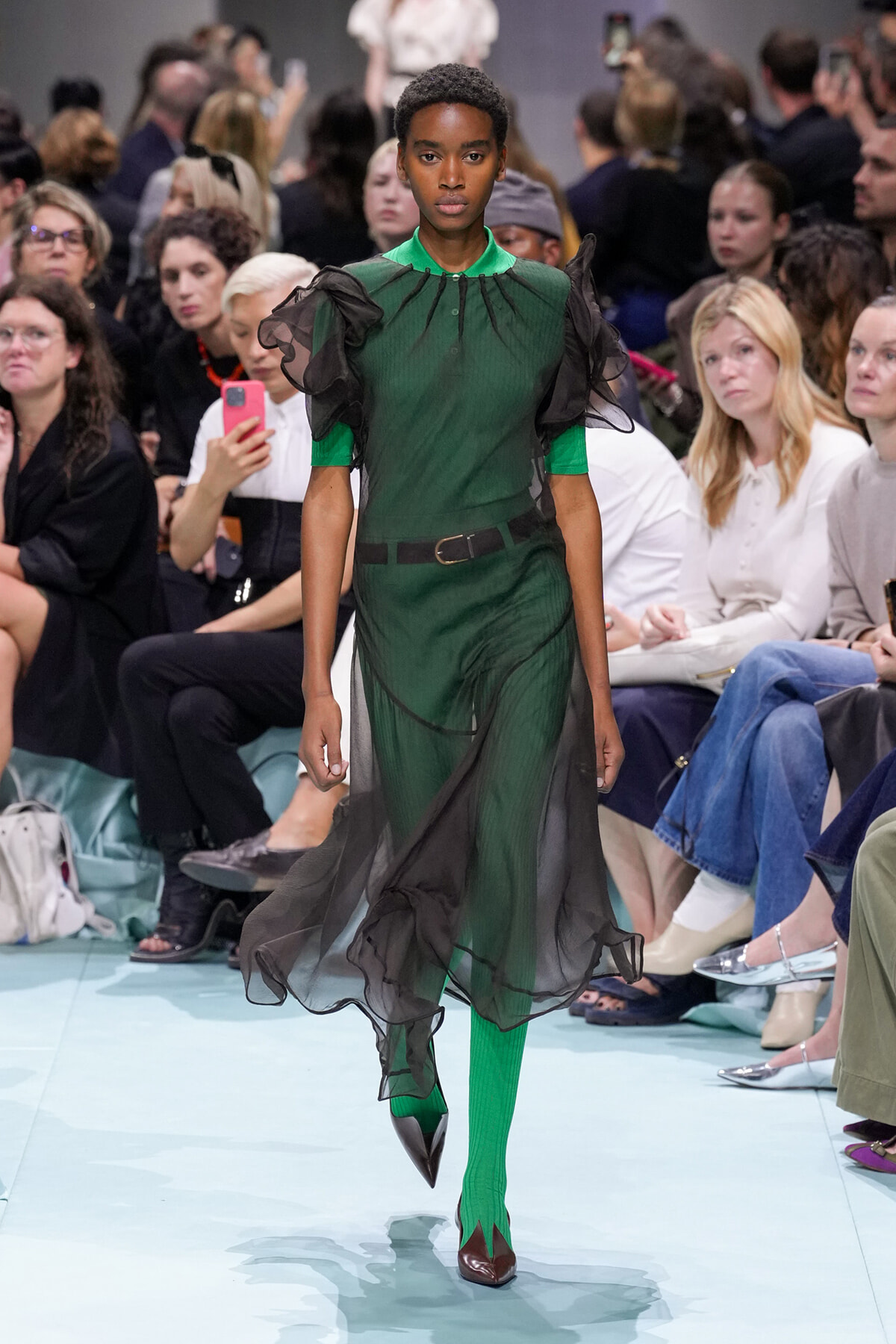 Model walking a runway in a green sheer dress with black organza overlay, matching green tights, burgundy heels, as the audience watches