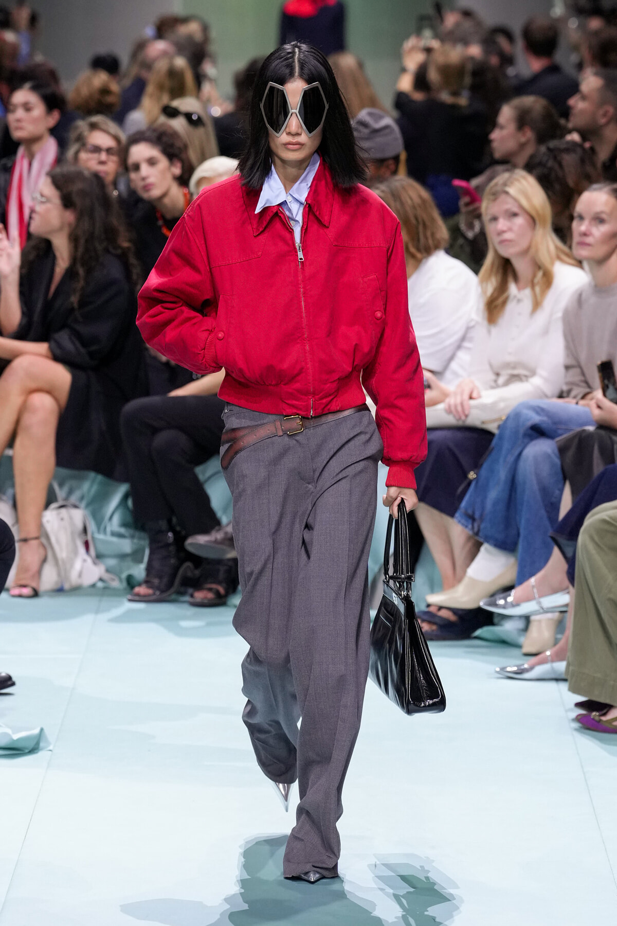 Model walking runway in a red bomber jacket, gray wide-leg trousers, oversized geometric sunglasses, and a black handbag.