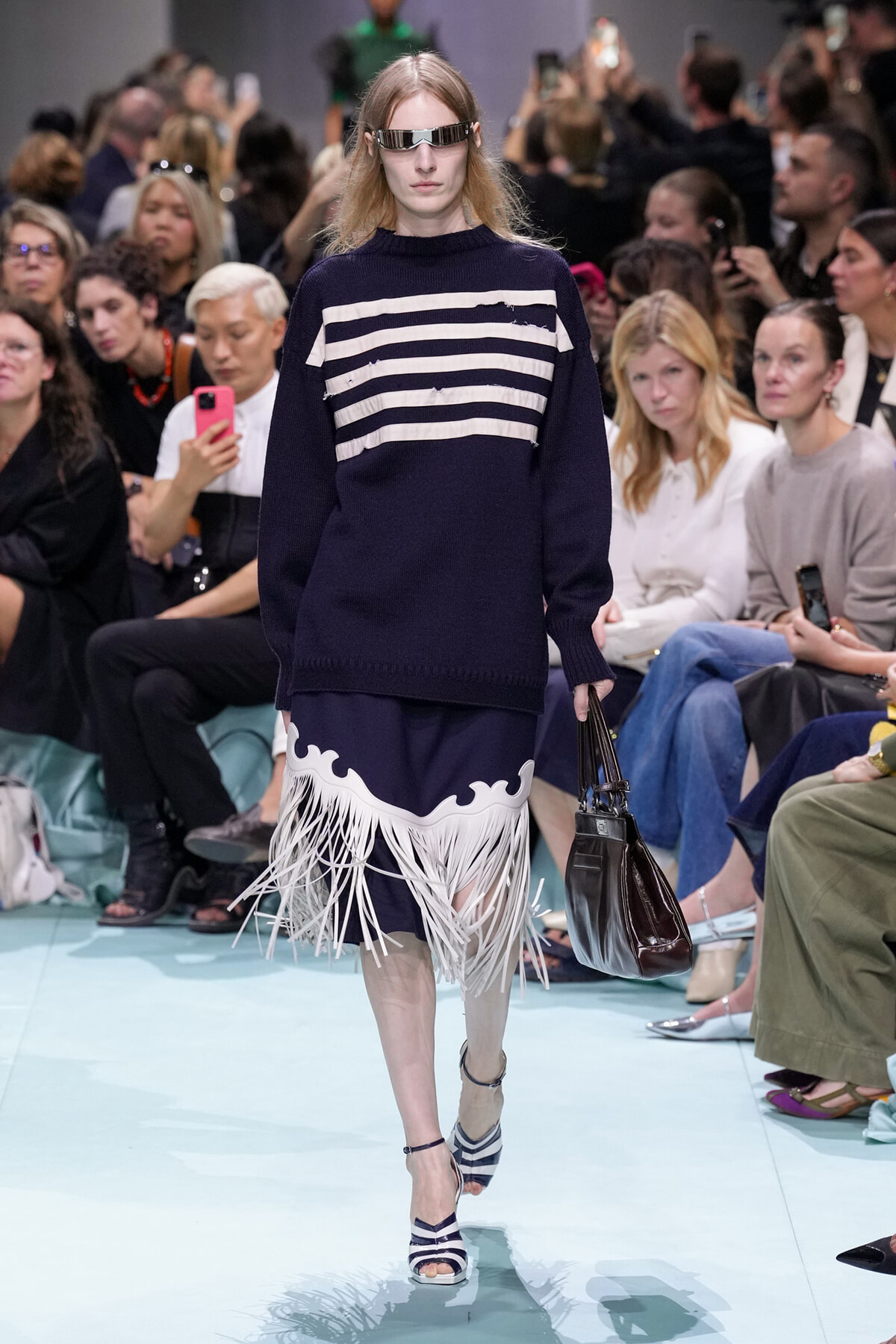 Model walks a runway in a navy sweater with white horizontal stripes and a white fringed hem over a navy skirt; she wears futuristic eyewear and carries a dark leather handbag.