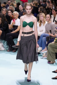 Model walks the runway in a green strapless bandeau top and a dark gray pleated midi skirt with a wide belt and metal hardware, audience in background.