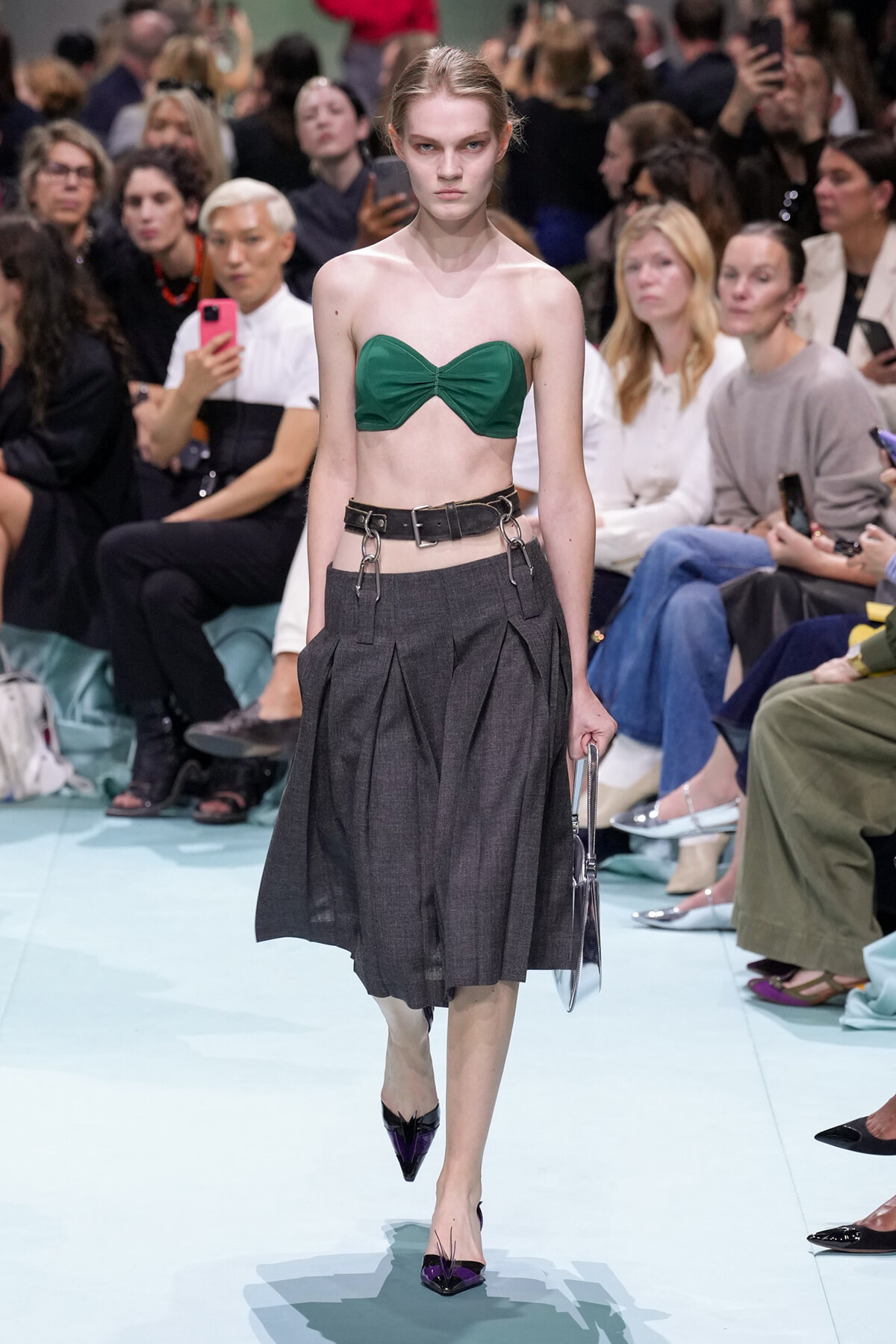 Model walks the runway in a green strapless bandeau top and a dark gray pleated midi skirt with a wide belt and metal hardware, audience in background.